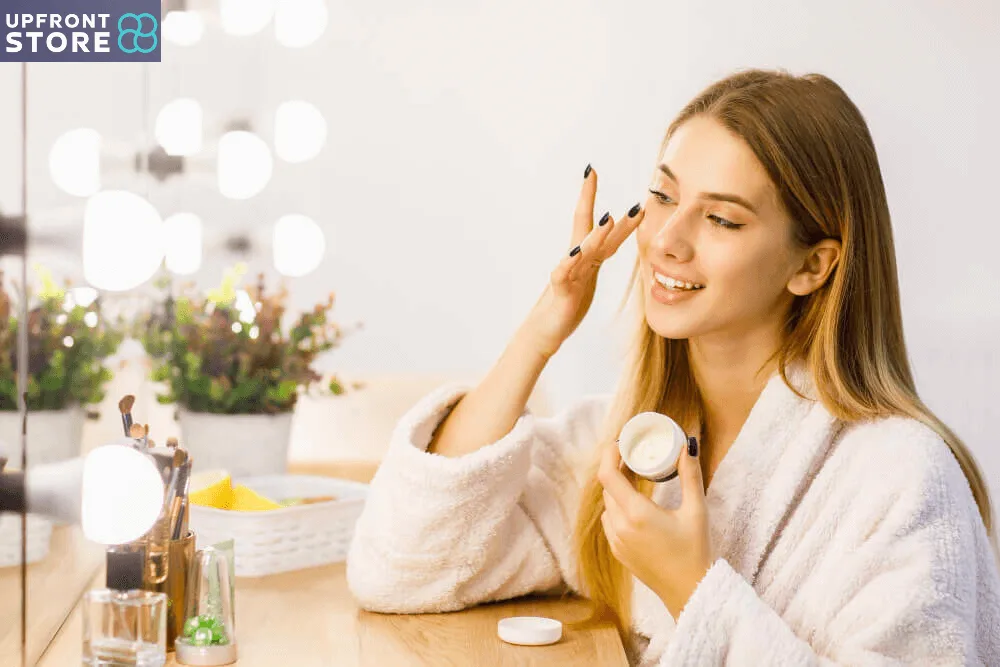 Smiling woman applying skincare cream in front of a mirror with glowing lights. Beauty and self-care routine featuring premium cosmetics from Upfront Store."